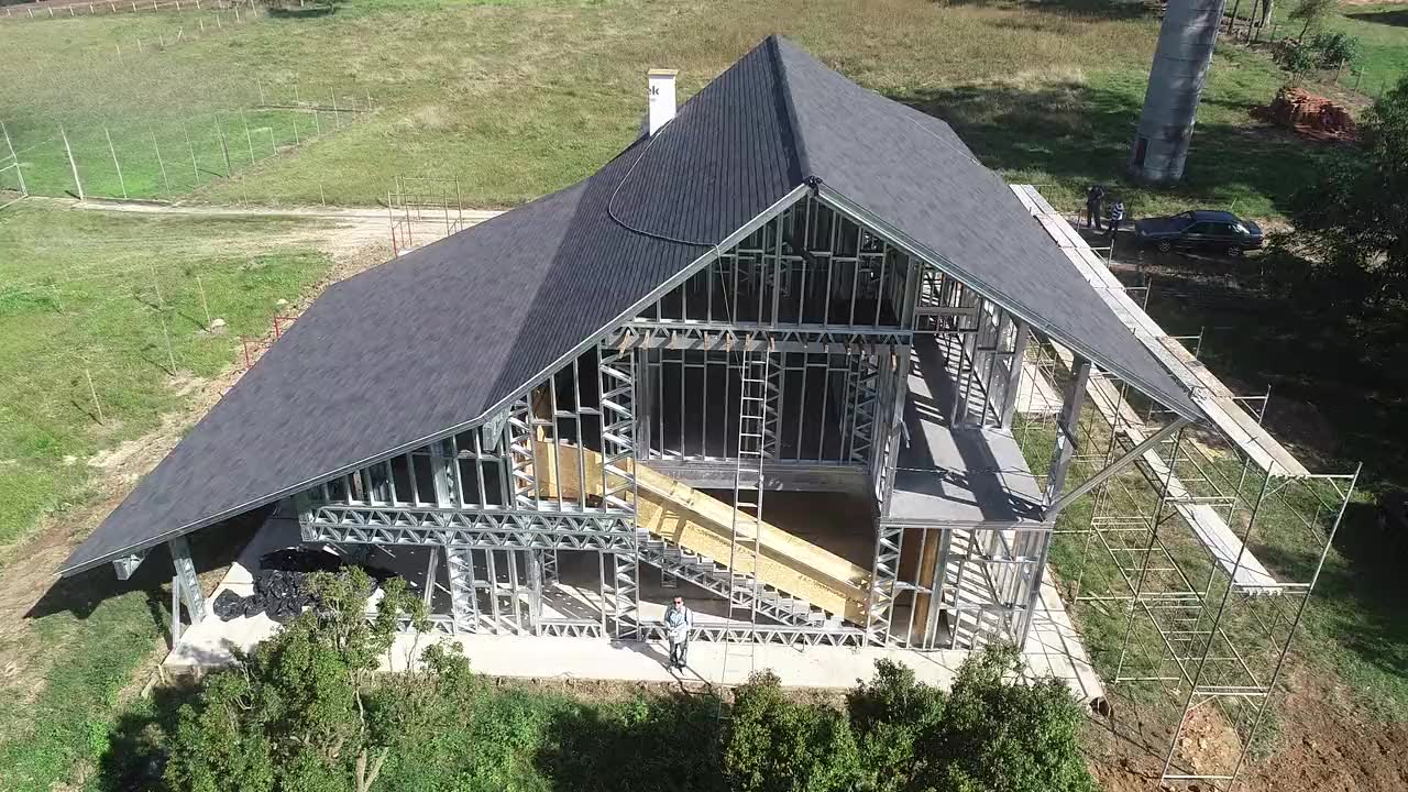Casa em light steel frame em obra, vista de drone: a estrutura de perfis de aço
            à vista debaixo da cobertura já telhada, rodeada de relva e árvores.